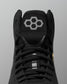 Close-up of a black high-top wrestling shoe showing the tongue with a gray geometric hexagon logo, black laces, and a small yellow accent near the side, set against a neutral gray background.