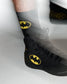 A close-up of a black high-top wrestling shoe with a yellow Batman logo on the side, worn with a black sock featuring a yellow Batman logo. The shoe has a textured knit upper and black laces, shown against a light gray background with a motion blur effect.