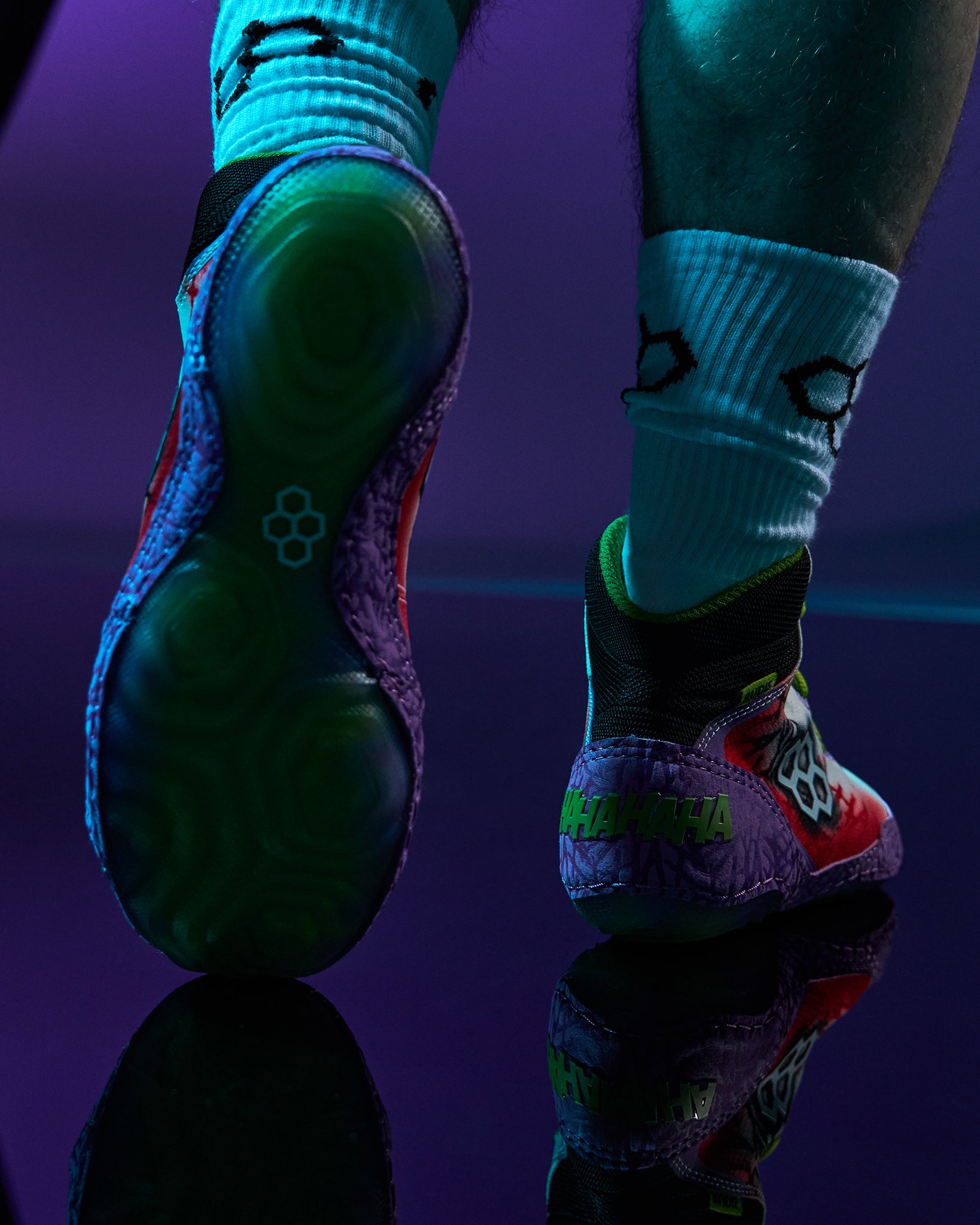 Close-up of a person's legs wearing colorful RUDIS wrestling shoes and white crew socks, with one shoe showing the sole and the other showing the side against a dark purple background.