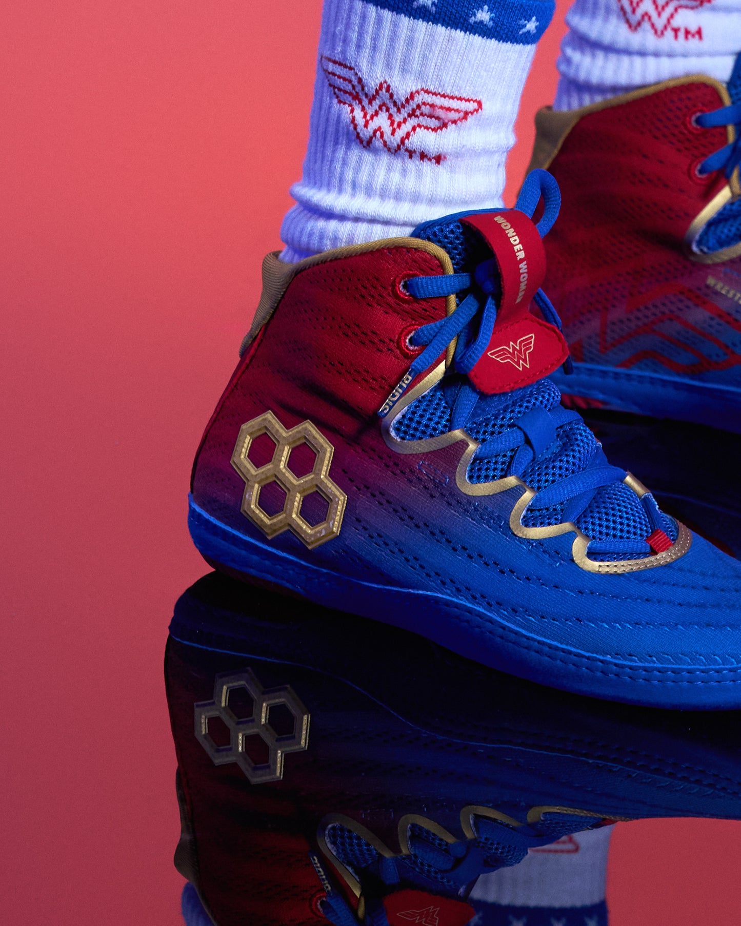 A close-up of red, blue, and gold high-top wrestling shoes with a reflective surface below, styled with white socks featuring a red and blue Wonder Woman logo, set against a solid red background.